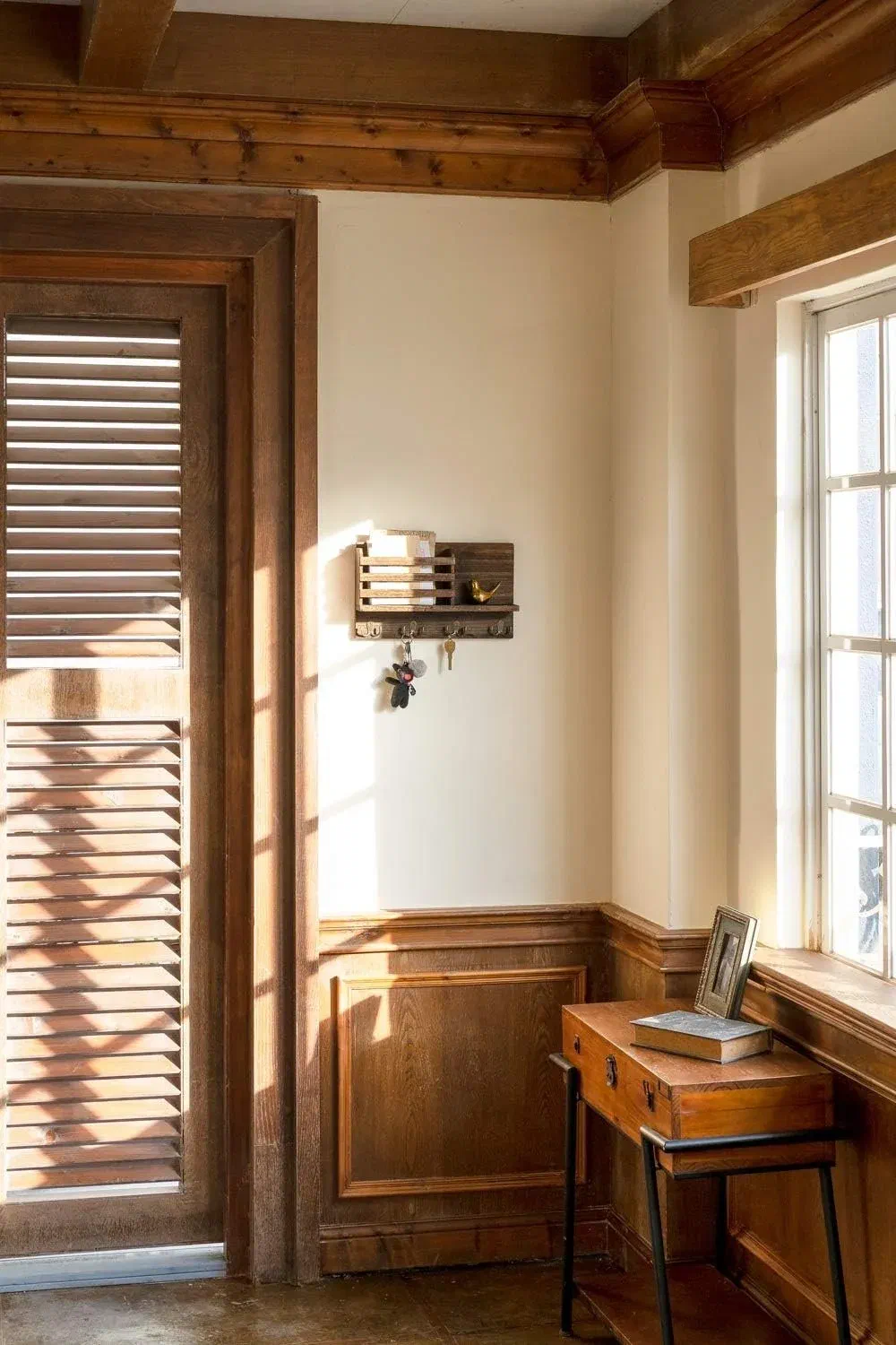 Wall-mounted wood mail and key organizer with keys hanging from its hooks, beside a sunlit entryway window