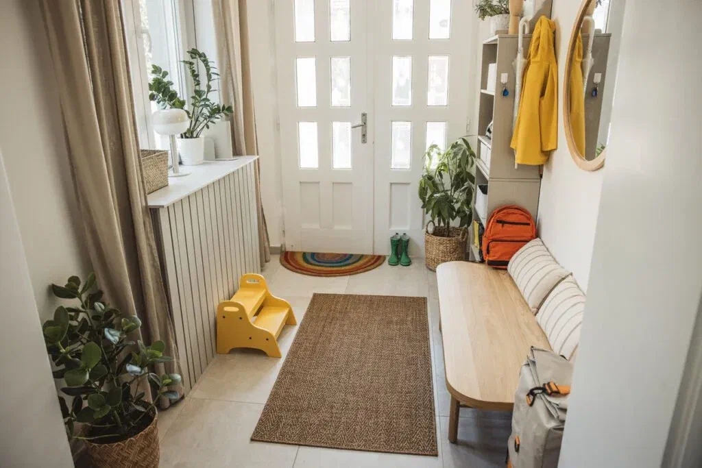 Bright entryway lined with potted faux plants beside a curtained window and bench, with a colorful mat by the front door
