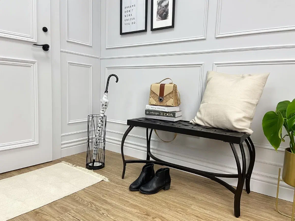 Wire umbrella stand beside a black bench topped with a cushion, a woven bag, and stacked books in a paneled entryway