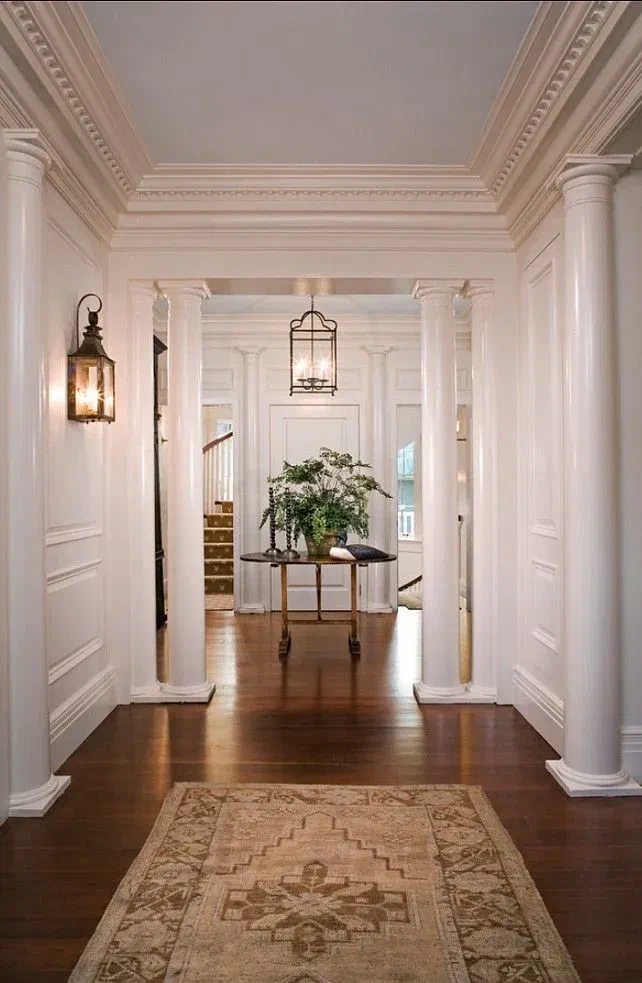 Grand columned foyer lit by wall lanterns and a hanging light fixture, with a round accent table and patterned rug