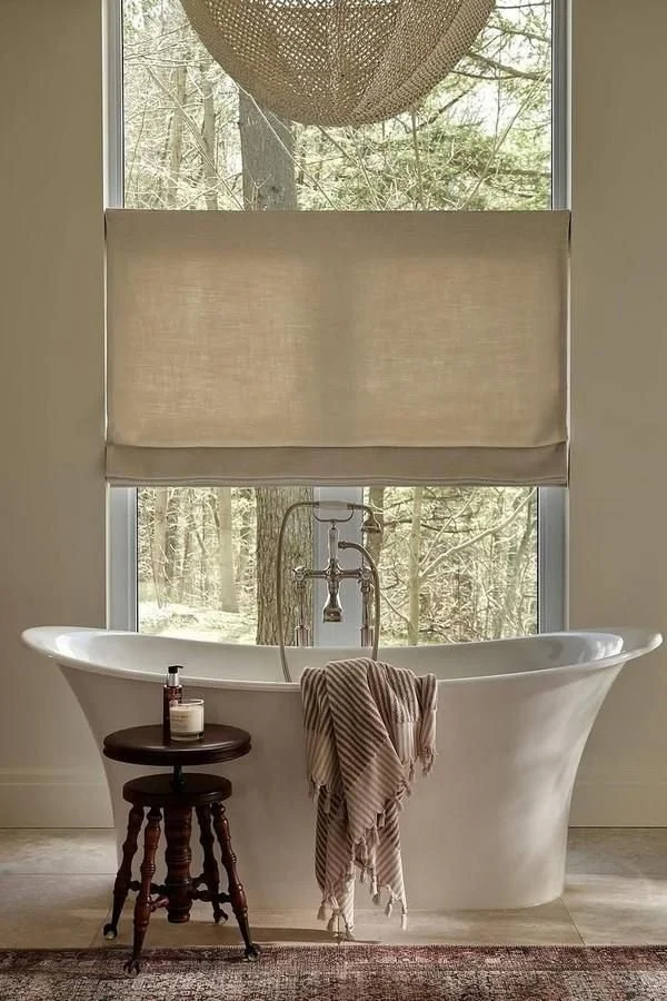 Freestanding white bathtub with a view of a forest, featuring a natural light-filtering blind.