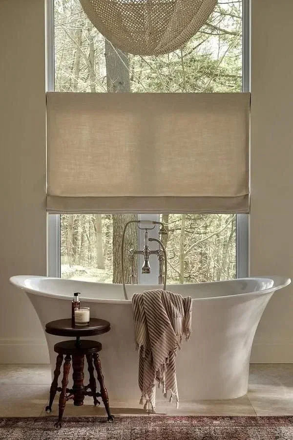 Freestanding white bathtub with a view of a forest, featuring a natural light-filtering blind.