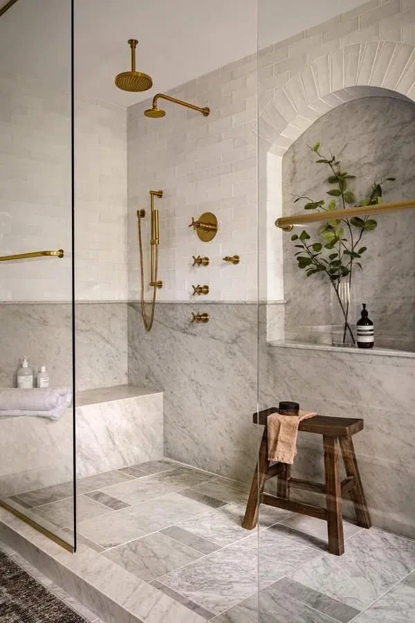 Spa-like shower with white subway tiles, marble accents, gold fixtures, and a wooden stool.