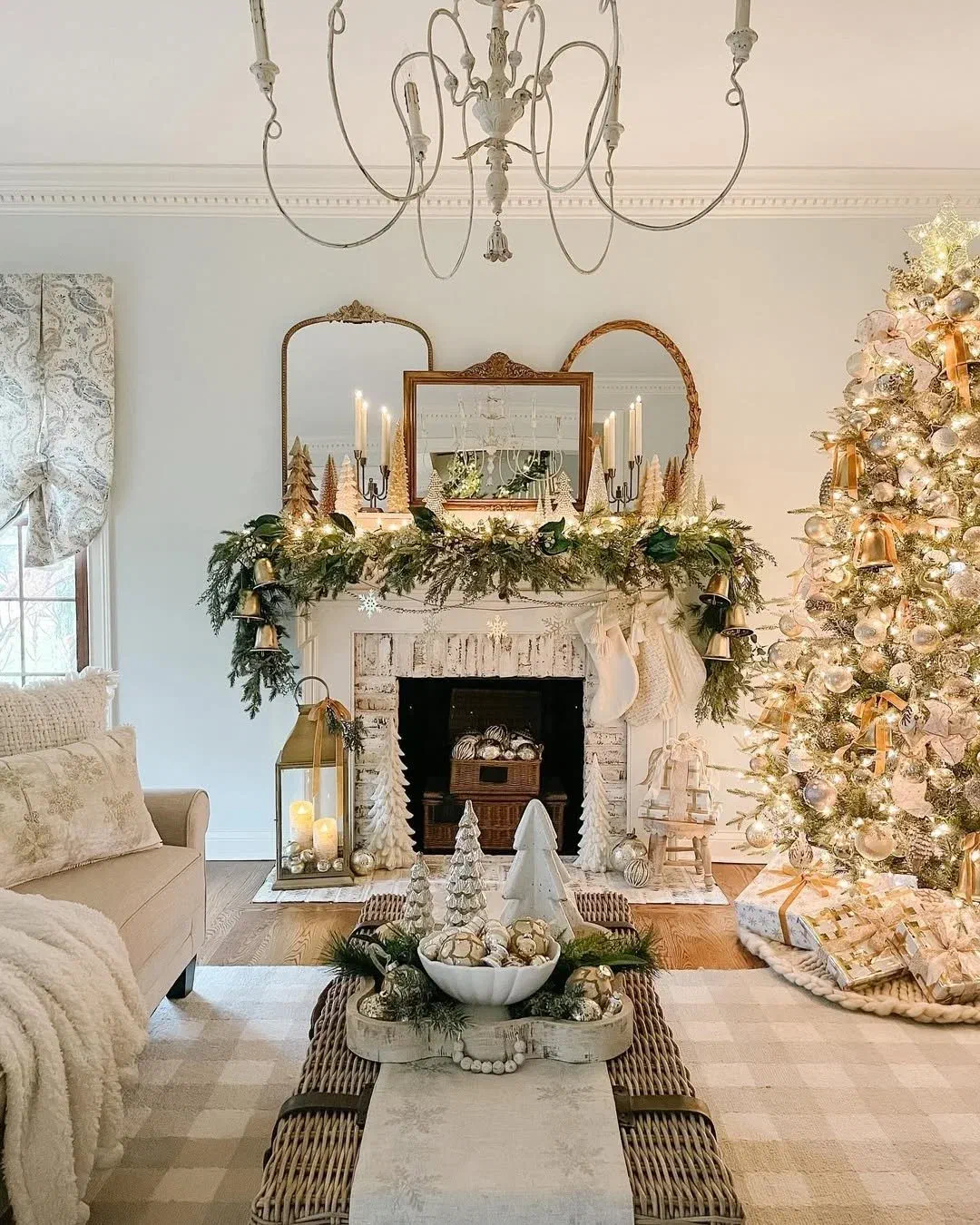 Neutral gold and cream living room with a lit Christmas tree beside a garland-draped mantel and antique mirrors