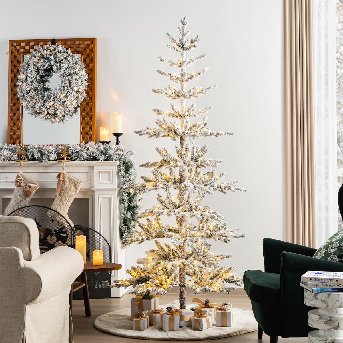 Snow-flocked pre-lit artificial Christmas tree glowing with warm white lights in a neutral living room, beside a flocked wreath and wrapped gifts