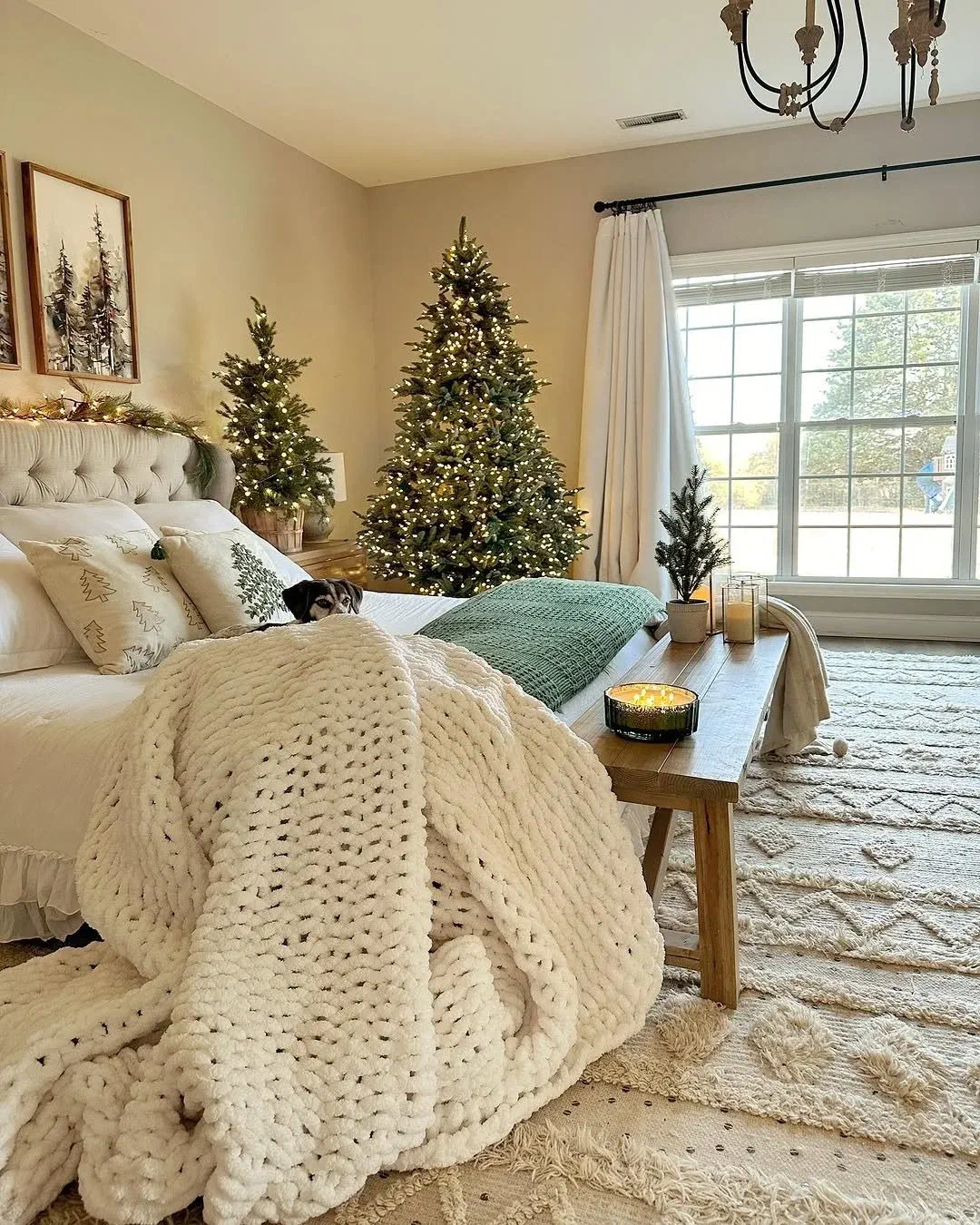 Cozy bedroom with two lit Christmas trees and a chunky knit throw blanket piled across the bed