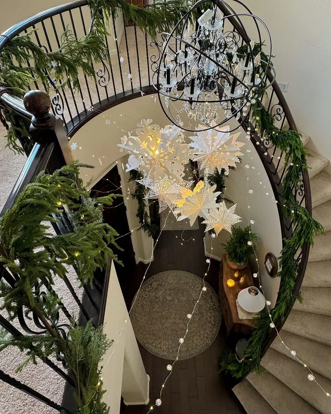 Curved staircase wrapped in green garland and fairy lights, with paper snowflakes hanging beneath a crystal chandelier