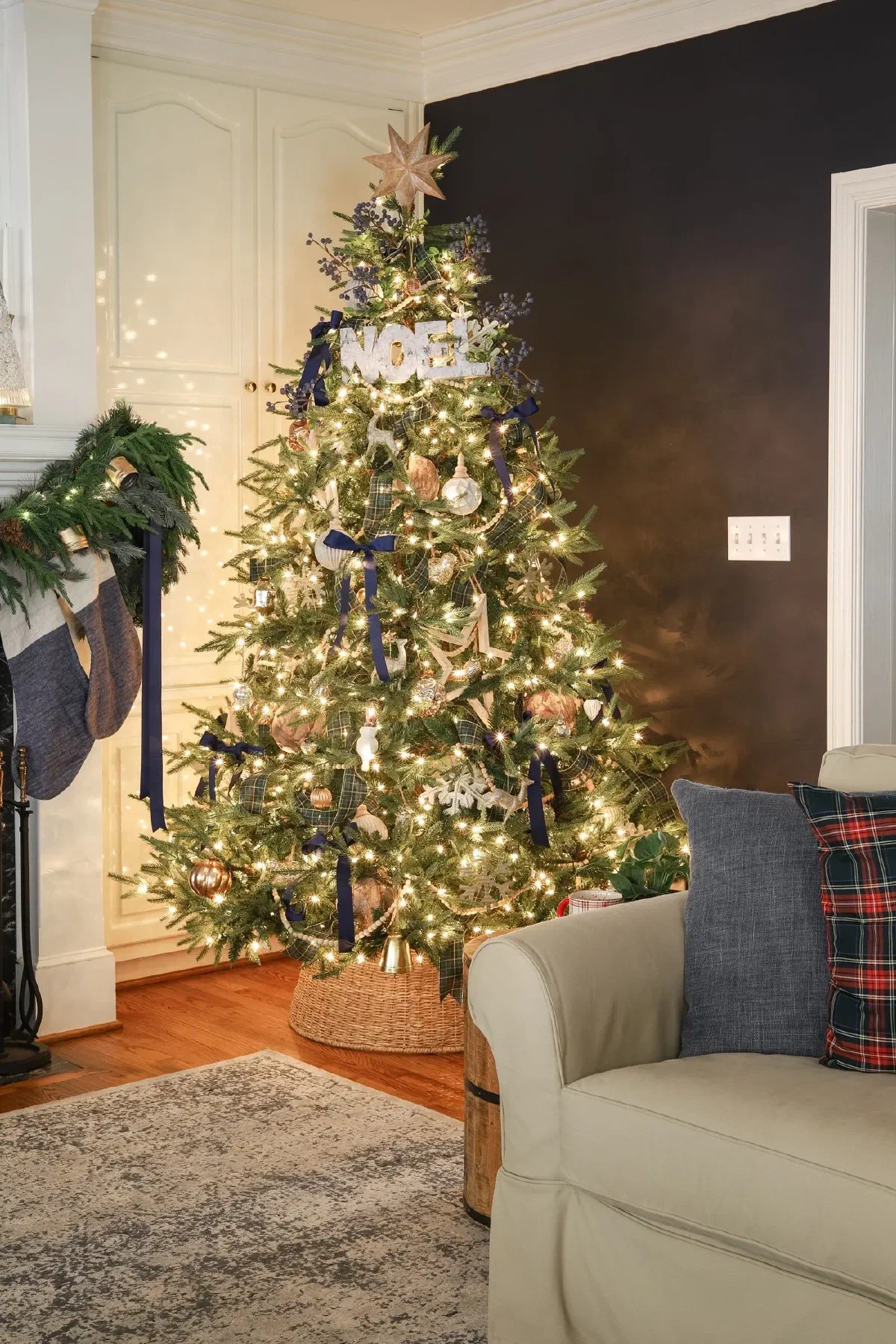 Christmas tree with a woven collar at its base, trimmed in navy ribbon, plaid, and a “NOEL” sign, beside a plaid-pillowed sofa