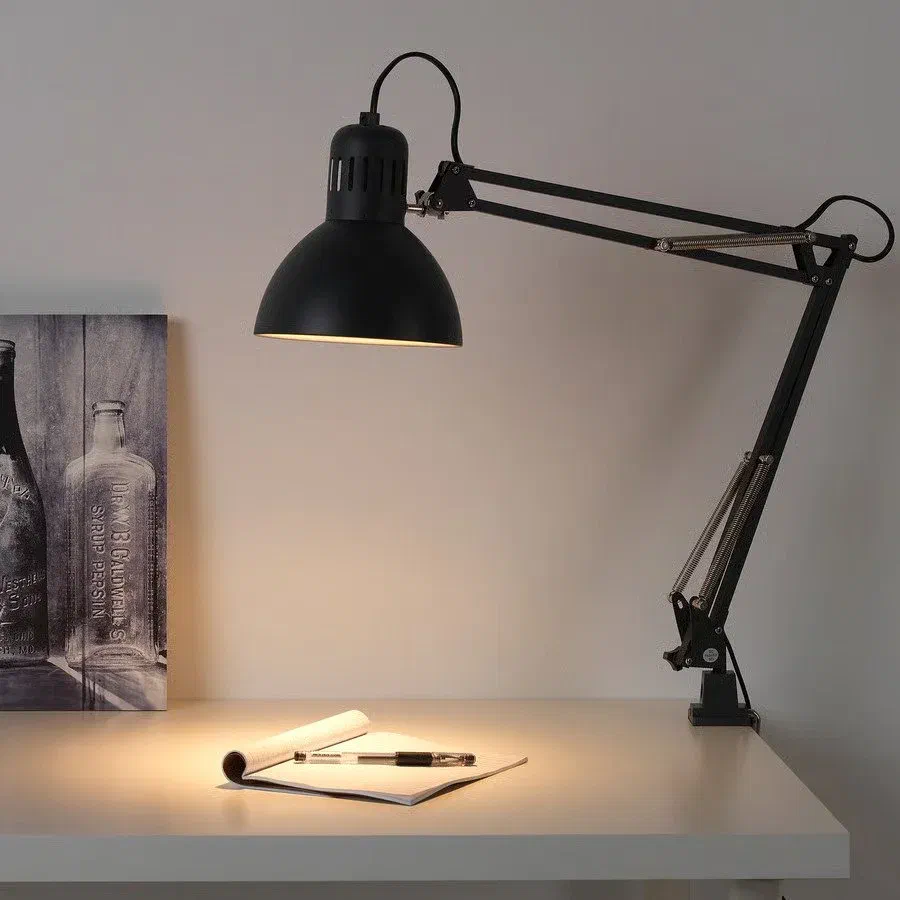 A black IKEA TERTIAL Work Lamp with a spring-loaded adjustable arm and a bell-shaped shade, illuminating a notebook and pen on a white desk. A framed black and white image is on the left.