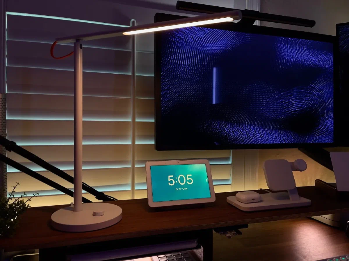 A white Xiaomi Mi LED Smart Desk Lamp with a slim, minimalist design and a flat light panel, placed on a wooden desk alongside a monitor displaying blue patterns and a smart home hub.