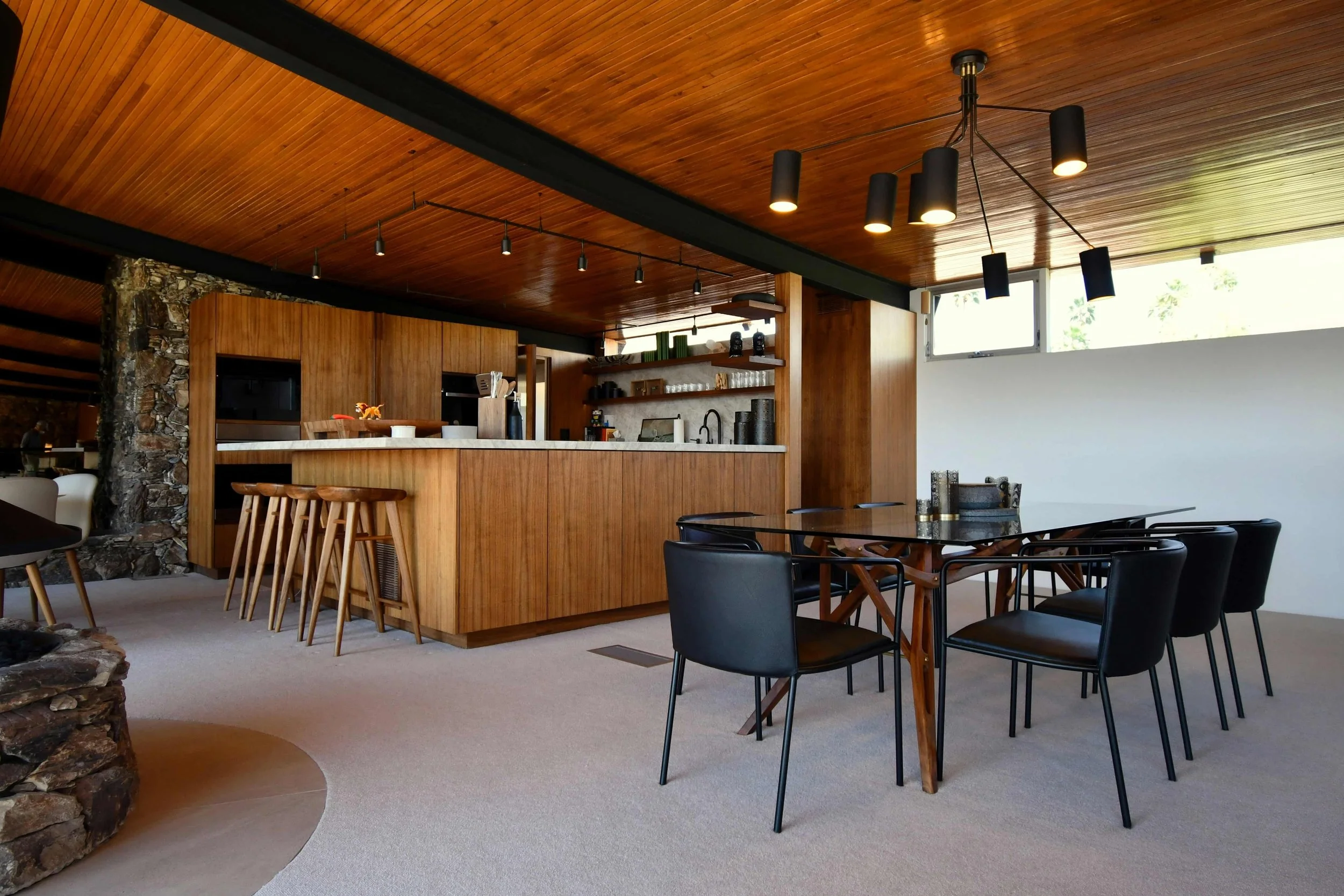 A spacious modern dining room with a large black multi-light chandelier hanging over a black dining table with matching chairs. The room has wood paneling on the ceiling and a stone accent wall.