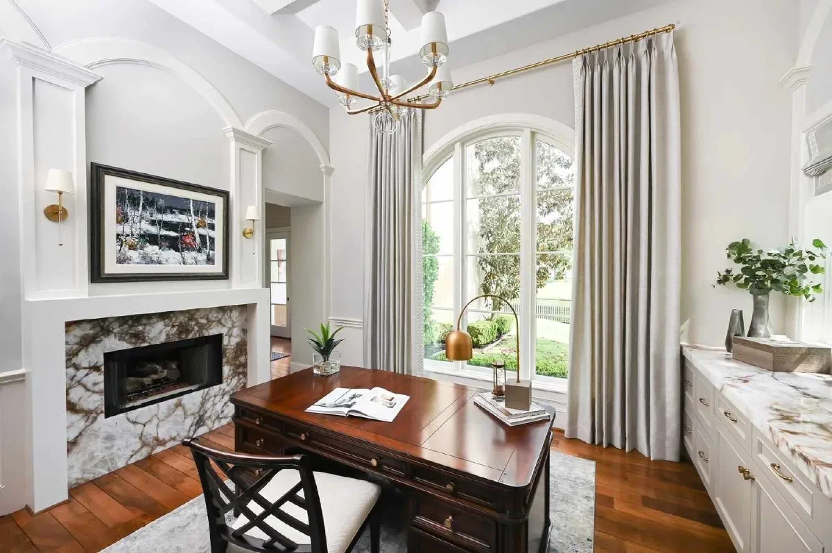Home office layout with large window for natural light, wooden executive desk, task lighting, light gray curtains, and fireplace. Ideal spot for a well-lit home office.