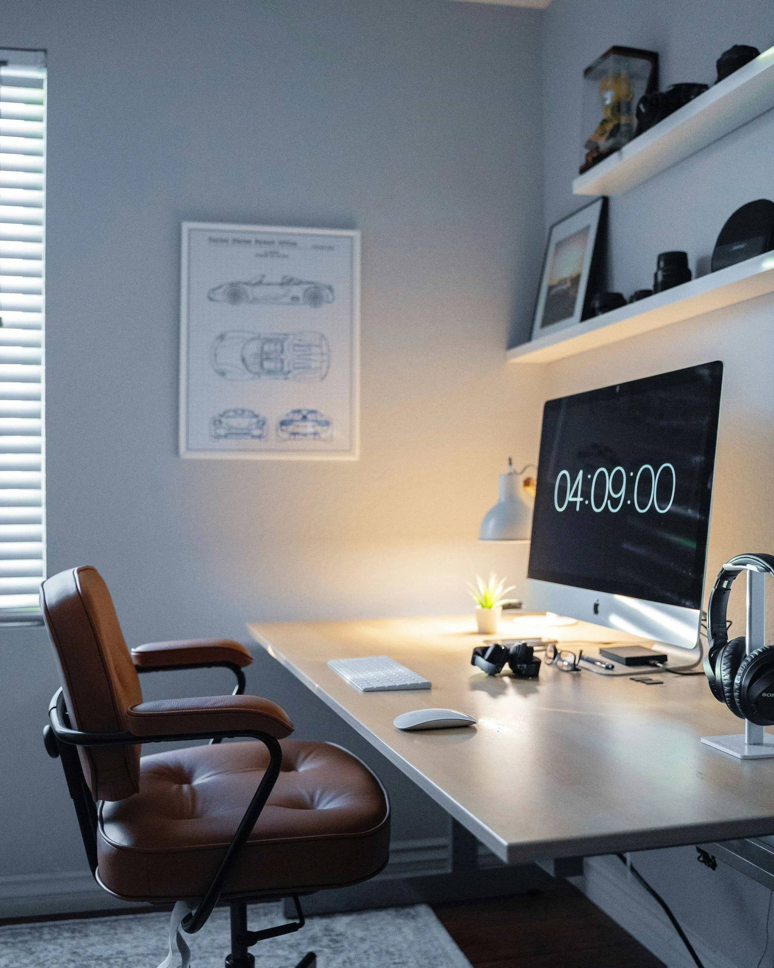 A home office desk with a large computer monitor displaying a clock, an ergonomic chair, and a framed blueprint of a car on a light blue wall, showing a productive and organized workspace.