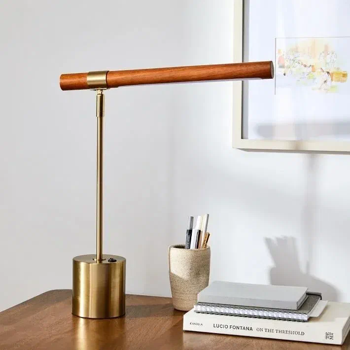 A modern and elegant desk lamp with a cylindrical brass base and a wooden horizontal light bar. The lamp sits on a wooden desk next to a book and a pen holder.