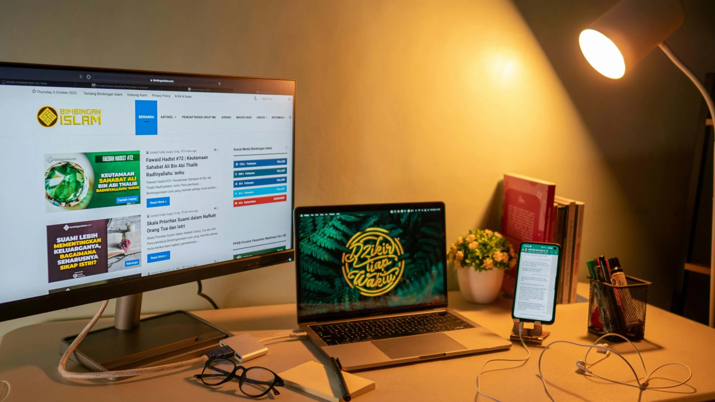 A home office setup with a dual-monitor display, a laptop, and a smartphone stand. The screens show a website and a leafy plant image, illuminated by the warm, directional light of a desk lamp.