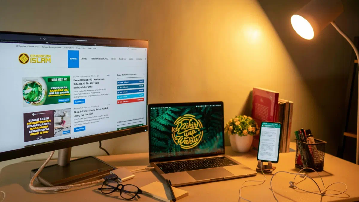 A home office setup with a dual-monitor display, a laptop, and a smartphone stand. The screens show a website and a leafy plant image, illuminated by the warm, directional light of a desk lamp.