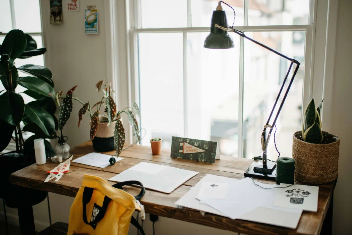 A creative and cozy home office desk with a large window in the background, featuring various plants, a yellow tote bag, and a large black adjustable desk lamp on a rustic wooden table.