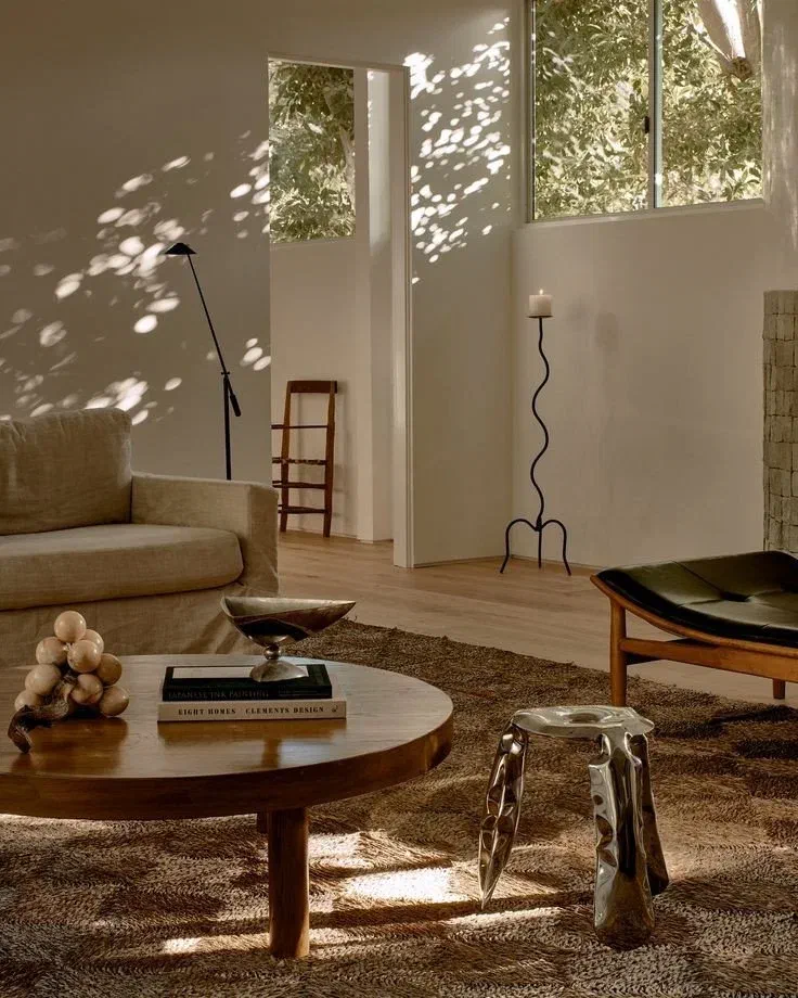 Sunlit minimalist living room with a linen sofa, wavy iron candle stand, and sculptural side tables on a patterned rug