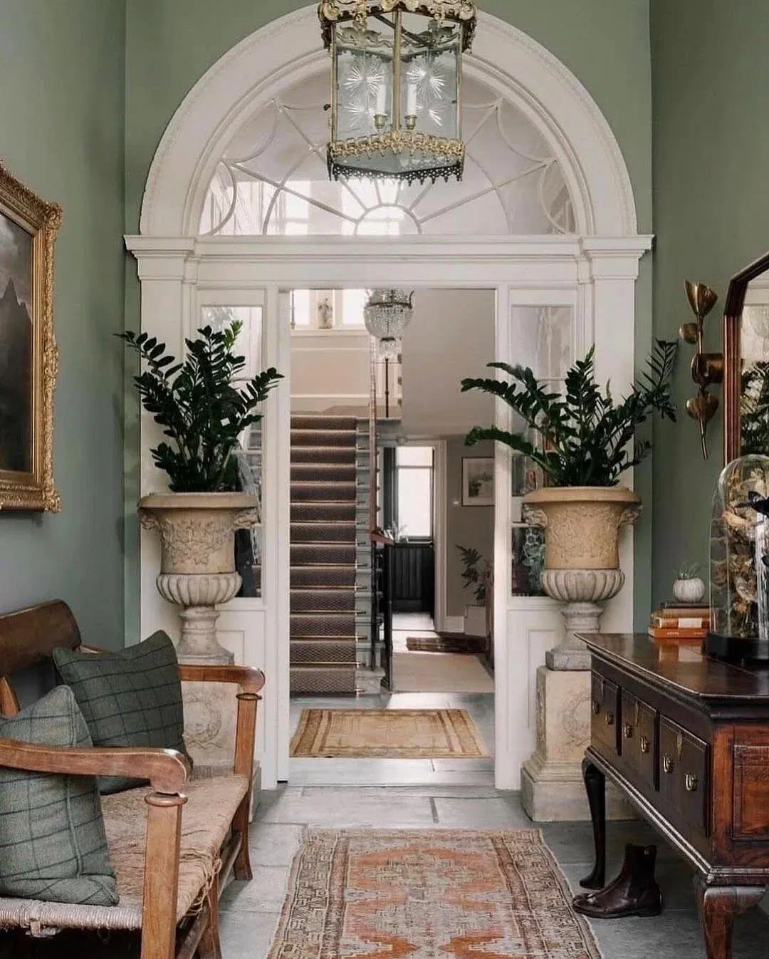 Sage green hallway with an arched doorway framed by tall stone urns of greenery, a plaid-cushioned bench, and antique furniture