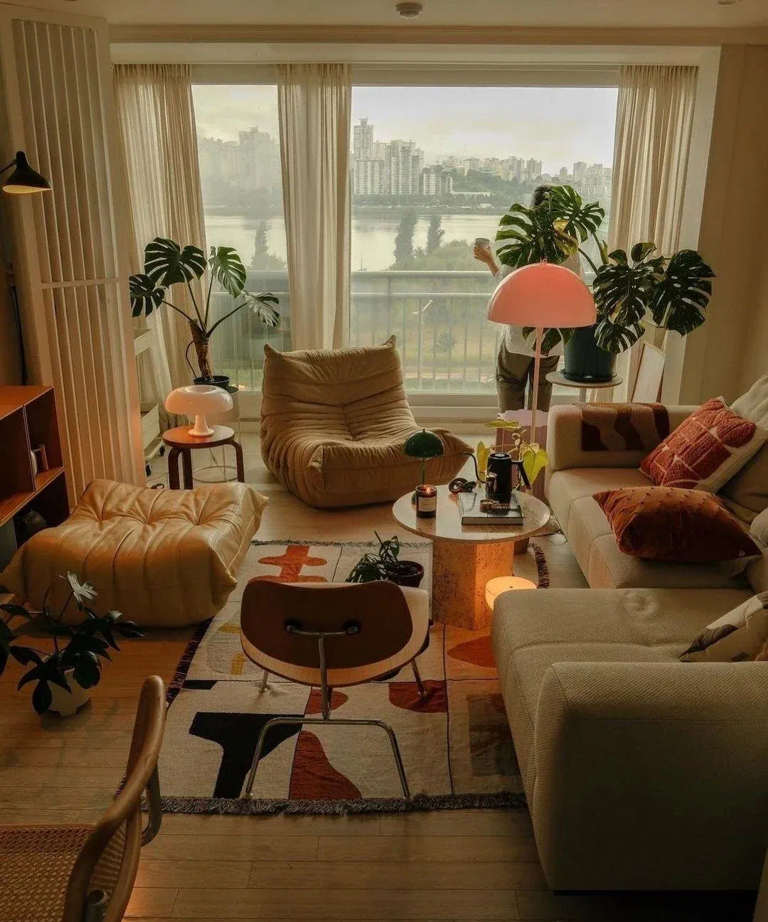 Mid-century living room with tufted caramel lounge chairs, a pink dome floor lamp, and monstera plants beside a city-view window